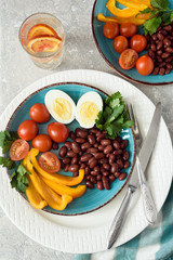 Healthy food, red beans, tomatoes, boiled egg, pepper with parsley in a plate with devices and a glass of water with orange on a gray background top view.