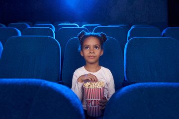 Pretty cute girl sitting with popcorn bucket in cinema.