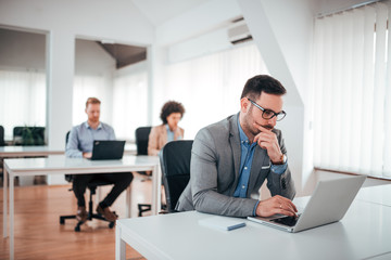 Handsome pensive businessman working on laptop in coworking office.