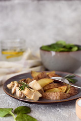Baked potatoes sliced ​​with chicken fillet in a creamy sauce in a plate with a fork on the background of a salad bowl with spinach and a glass of water and lemon.