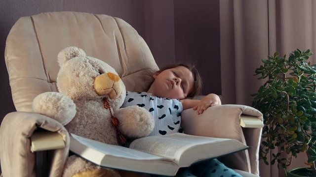 Little girl sleeping on the chair with big book hugging toy teddy bear
