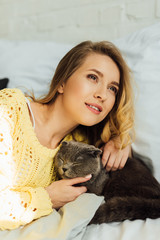 beautiful smiling girl in knitted sweater lying in bed and hugging scottish fold cat