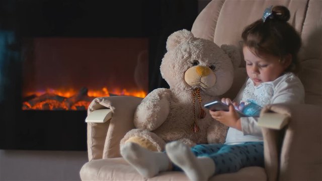 Little girl sitting on the chair with smartphone hugging toy teddy bear at night. Home furnishings with fireplace
