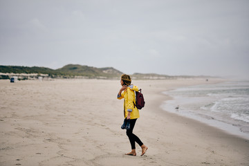 Young woman wearing yellow rain jacket strolling along North Sea Shore barefoot carrying her shoes...