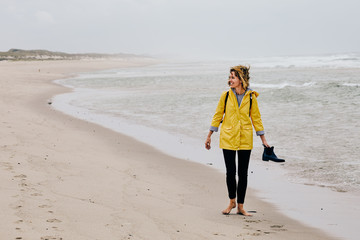 Young woman wearing yellow rain jacket strolling along North Sea Shore barefoot carrying her shoes in the hand - obrazy, fototapety, plakaty