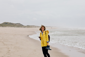 Young woman wearing yellow rain jacket strolling along North Sea Shore barefoot carrying her shoes...