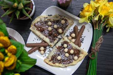 Homemade pancakes with chocolate, chocolate crumbs and candies on white plate, and flowers on wooden table