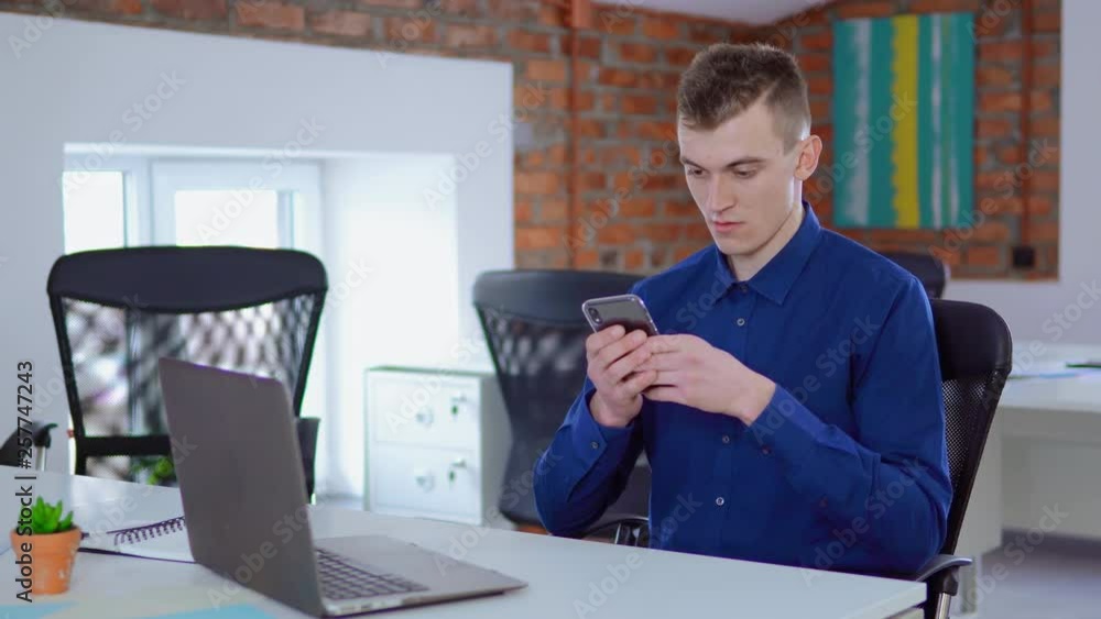 Poster young worker in startup company using smartphone texting message or checking email sitting at the white desk in loft office businessman at work daytime