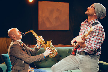Two Caucasian great musicians playing bass guitar and saxophone in home studio.