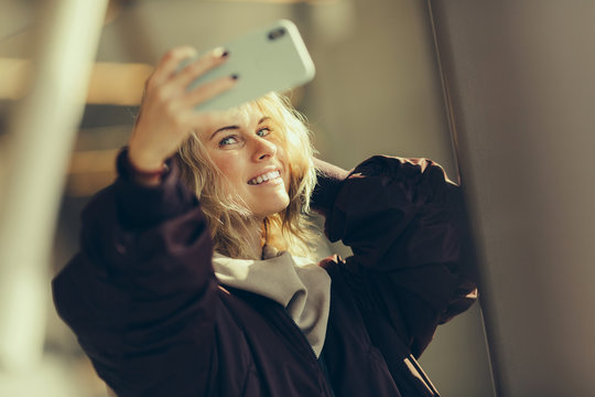 Young happy blonde in jacket makes selfie against background of window