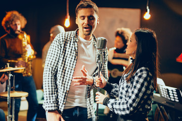 Singers practicing for duet. In background band playing. Home studio interior.