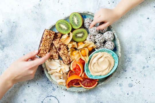 Mother sharing healthy vegan dessert snacks with toddler child. Healthy sweets for children. Protein granola bars, homemade raw energy balls, cashew butter, toasted coconut chips, fruits platter