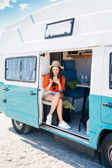 Young woman sitting in her camper van smoking and using smartphone