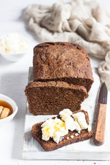 Slice of dark rye bread with fresh farm cheese and honey on a white wooden cutting board.