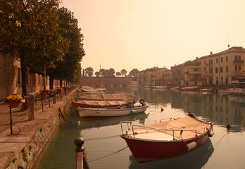 Scenic view of a canal with moored boats and a tree line on the bank at sunset, Peschiera del Garda, Veneto, Italy
