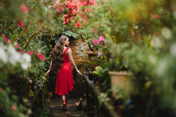 Young beautiful caucasian woman in glass greenhouse among colorful azalea flowers. Art portrait of a long-haired girl wearing a romantic red dress.