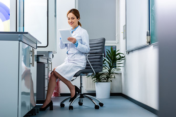 Smiling chemistry teacher in white coat using digital tablet while sitting in armchair in classroom