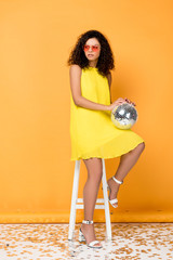 attractive curly african american woman holding disco ball while sitting on chair on orange