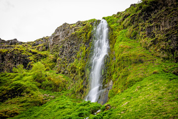 Obraz premium Small waterfall by Seljalandsfoss, Iceland with white water falling off cliff top long exposure in green mossy summer rocky landscape