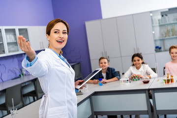 Smiling chemistry teacher in white coat holding digital tablet and pointing with hand