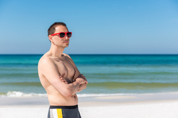 Young handsome attractive fit muscular man hipster millennial on beach during sunny day with red sunglasses in Florida panhandle ocean looking to side