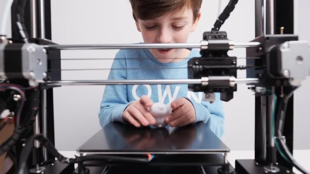 Printing on 3D printer figurines of toys from white plastic close-up. Boy happily and emotionally takes the finished printed figure from working platform of printer and goes into the play