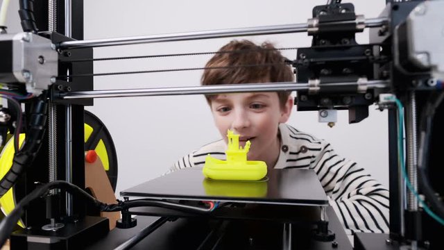 Printing on 3D printer figurines of toys from yellow plastic close-up. Boy happily and emotionally takes the finished printed figure from the working platform of the printer and goes into the play