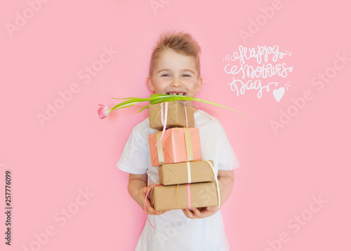 A little boy holds gifts and a postcard for mom. Mother's Day to celebrate. Pink background.