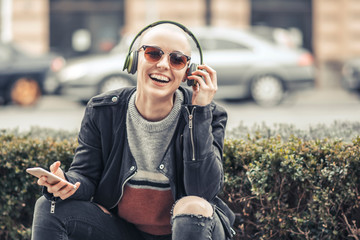 Young modern woman with bald hairstyle listening to her favorite music over her big green headphones, outdoor urban scene