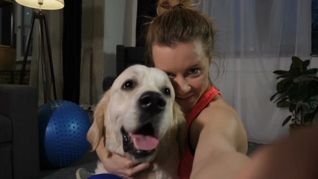 Closeup portrait of pretty fit female owner embracing big golden retriever dog and smiling on camera of smart phone in domestic room. Happy friends proudly posing for selfie photo after home workout