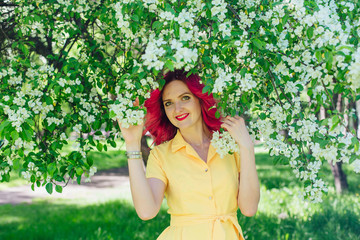 Beautiful and young woman with bright red hair and ref lips standing next to a blooming apple tree...