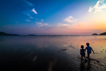 Far-away view of a muddy shore at low tide with children with blue and orange gradient sky and wispy clouds