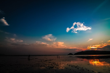 Far-away view of a muddy shore with children running and blue and orange gradient sky with dramatic sunset