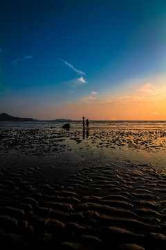 Far-away view of a muddy shore at low tide with children with blue and orange gradient sky and wispy clouds