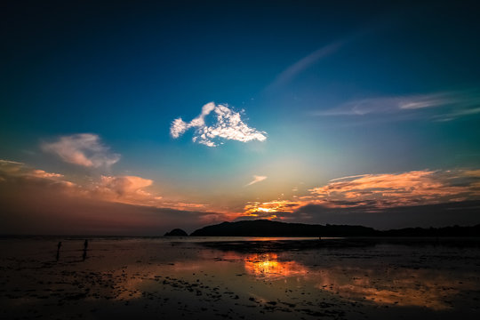 Muddy shore with two children walking with blue, white and orange gradient sky and wispy clouds