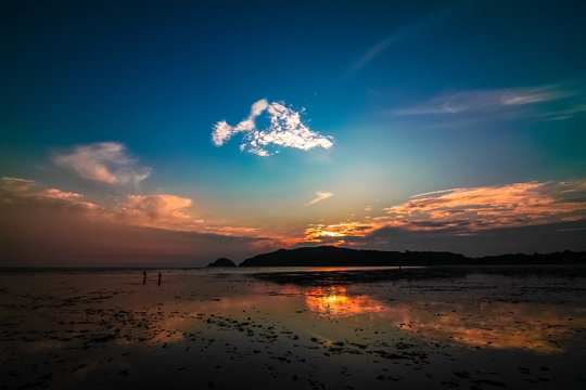 Far-away view of a muddy shore with children running and blue and orange gradient sky with dramatic sunset