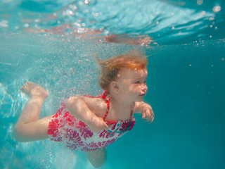 Obraz na płótnie Canvas Smiling baby girl in cute modern dress diving underwater in blue swimming pool. Active lifestyle, child swimming lesson with parents. Water sports activity during family summer vacation in resort