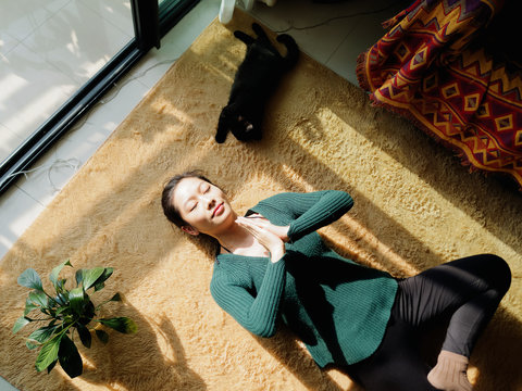 Pretty Chinese young woman meditating at home, lying on floor with her black cat in sun light, exercise, Lotus pose, prayer position, namaste, working out, Feeling peace and wellness concept.
