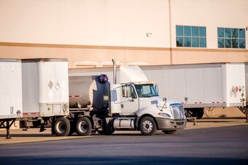 White big rig day cab semi truck with tank semi trailer standing in warehouse dock with another loading semi trailers