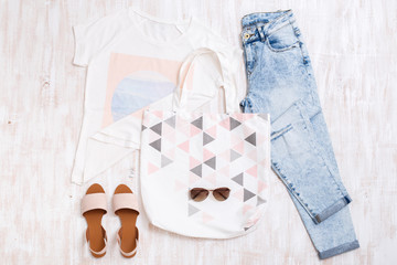 White t-shirt with print, light blue ripped boyfriend jeans, beige sandals, sunglasses, cotton textile bag on white wooden background. Overhead view of woman's casual outfits. Flat lay, top view.