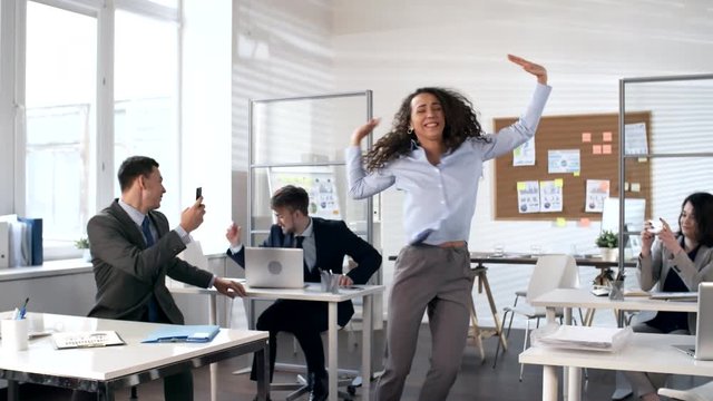 Young energetic businesswoman dancing with excitement in the center of open space office while her colleagues recording videos with smartphones and man throwing up documents in the air
