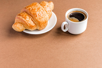 Tasty breackfast. French croissant served on white plate and cup of black coffee or espresso on brown background. Copy space.
