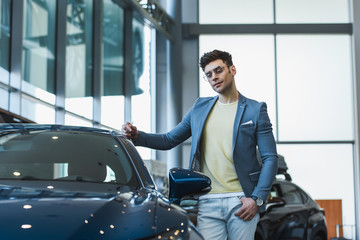 pensive man in glasses standing with hand in pocket near automobile in car showroom