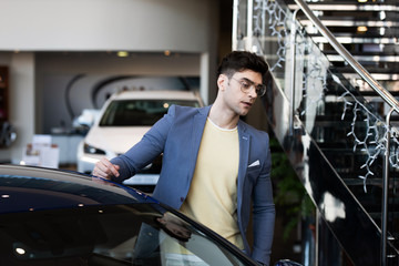 Fototapeta na wymiar handsome customer in glasses standing near automobile in car showroom