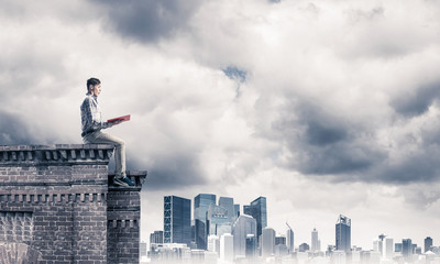 Man on roof edge reading book and cityscape at background