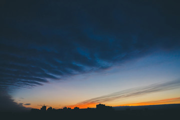 Cityscape with wonderful varicolored vivid dawn. Amazing dramatic blue cloud sky above dark silhouettes of city building roofs. Atmospheric background of orange sunrise in overcast weather. Copy space