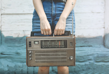 Hipster girl in denim jeans shorts is holding retro radio in hands. Style concept. Vintage music on wooden background. Modern technologies outdoors.