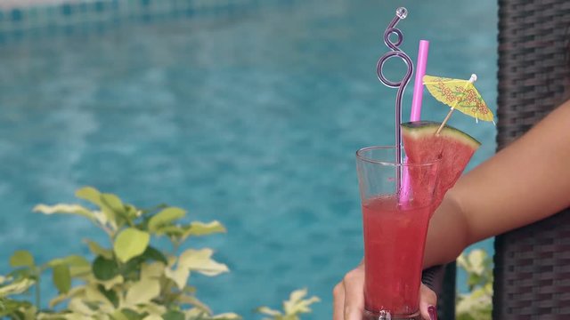 girl holds tall glass with watermelon cocktail and curvy purple stick against blue swimming pool