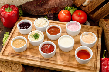 Assorted sauces: ketchup, perigueux, lesser, soy sauce, pesto, berry sauce, Dijon, bechamel in white sauce boats on a wooden background with vegetables. Top view. Close-up. Space