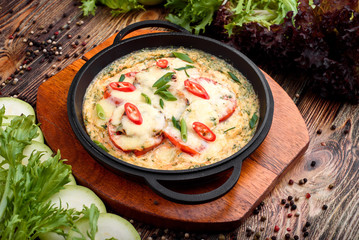 Omelet with cheese, tomatoes and chilli peppers in a cast iron pan on a wooden board on a dark wooden background, decorated with lettuce and fresh vegetables. Close-up. Space